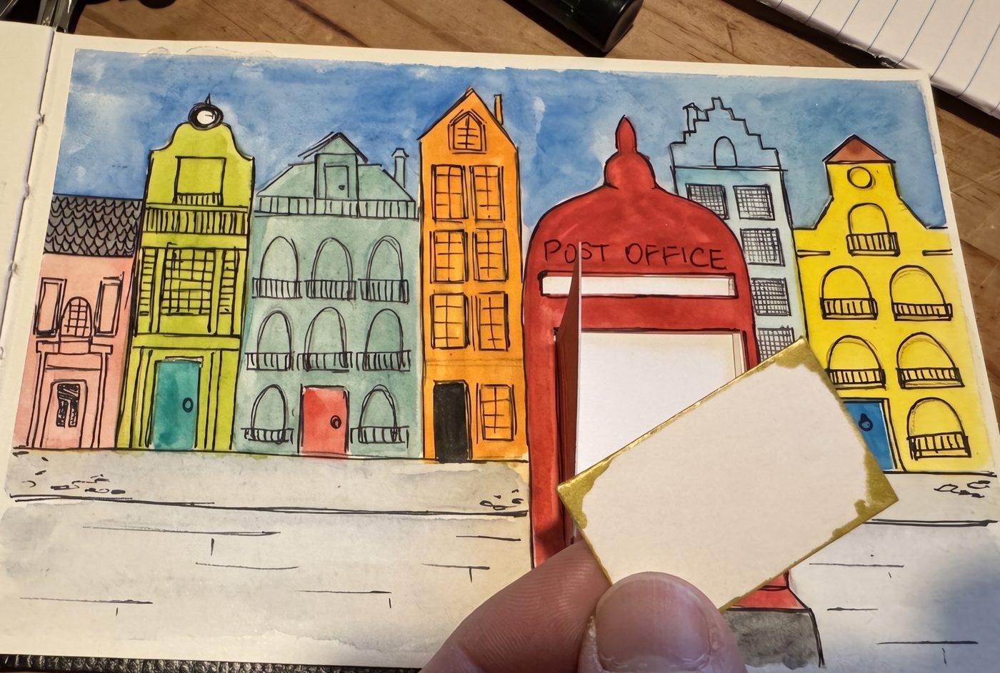 A watercolour Letterbox Street painting with a row of colourful townhouses and a red post office letterbox, with a tiny letter ready to post through the slot