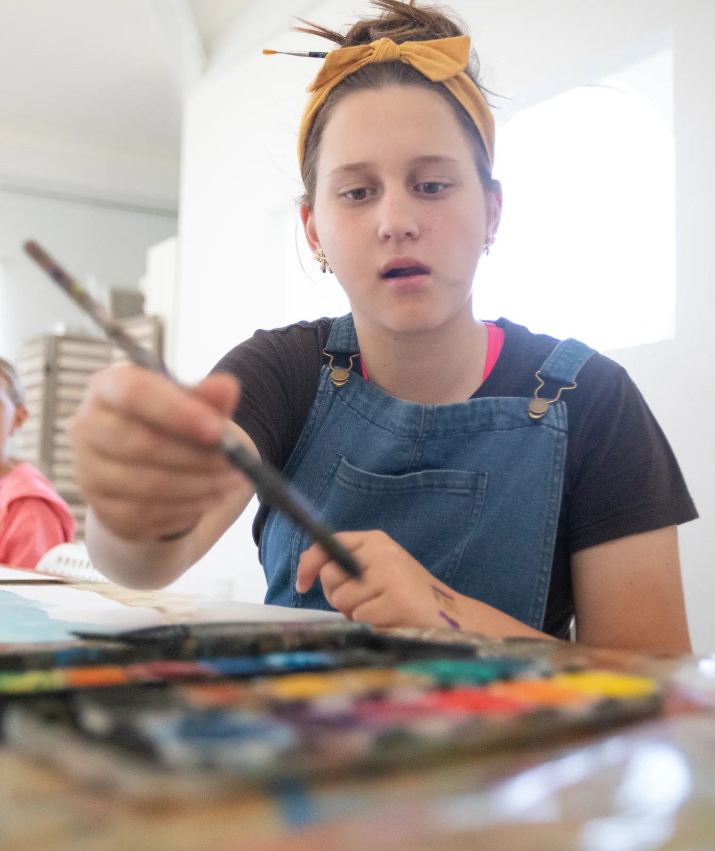A mini-artist loading a brush from a watercolour palette at the studio table, with another mini-artist painting behind her