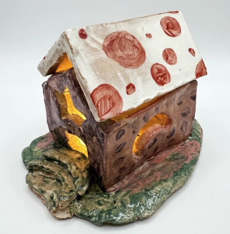 A hand-built ceramic clay house with a spotted roof, glowing from inside