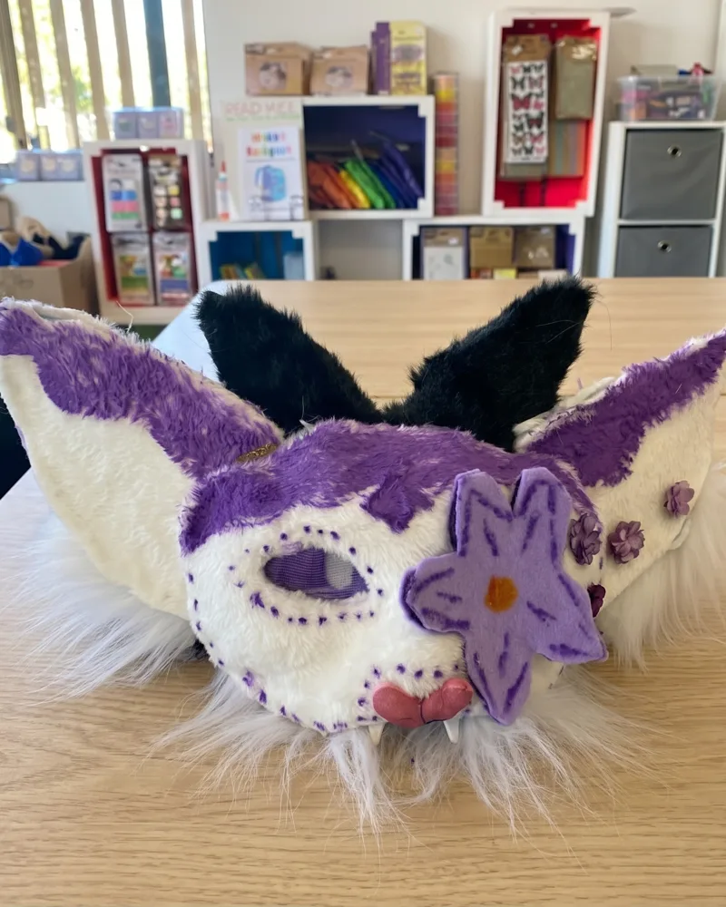 A furry white and purple creature half-mask with pointed ears and a felt flower