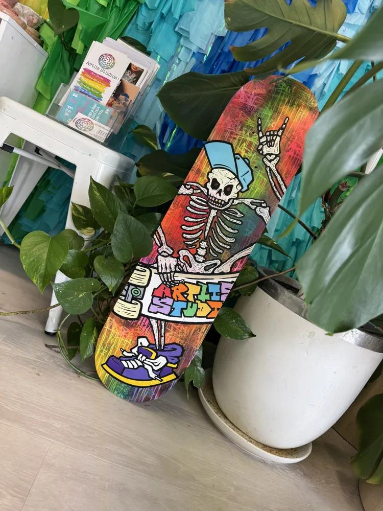 A skateboard deck painted with a rainbow background and a skeleton in a blue cap